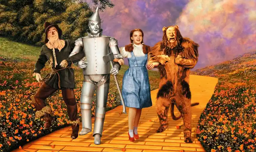 Auf dem Bild abgebildet sind von links nach rechts aus dem Film Der Zauberer von Oz von 1939 die Vogelscheuche, der Blechmann, Dorothy und der Löwe wie sie, die Arme ineinander verschlungen, auf der gelben Backsteinstraße in Richtung Smaragdstadt wandern.