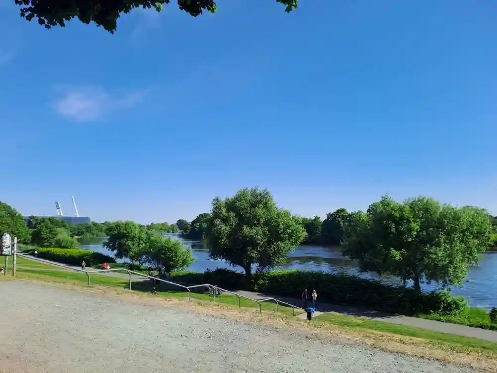 Ein Flaneur am Weserdeich blickt auf die Stadt, wo grüne Deiche den urbanen Asphalt durchziehen und das Weser-Stadion als Mahnung an vergangene Zeiten in den Himmel ragt. Regen, Reflexionen und stille Momente verweben Natur, Geschichte und städtisches Leben.